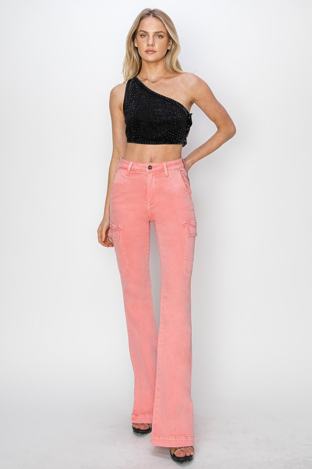 A person wearing RISEN Full Size High Rise Side Slit Cargo Bootcut Jeans, featuring a pink flared design with front pockets, stands against a plain background.