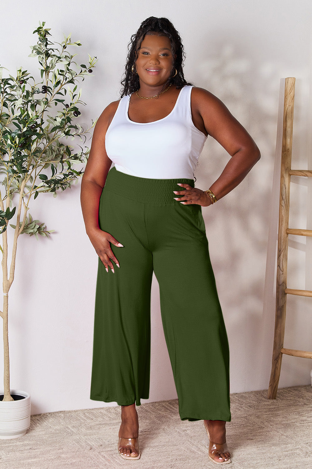 A curve model is wearing the Double Take Full Size Smocked Wide Waistband Wide Leg Pants in gray with hands in pockets, paired with white sneakers.