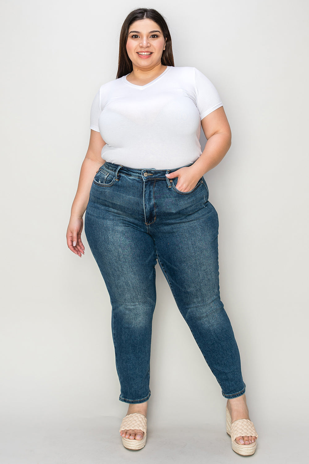 Person wearing Judy Blue Full Size Tummy Control High Waist Slim Jeans in blue and a white shirt, with their hands in their pockets. They are standing indoors on a polished concrete floor with a wooden cabinet and plant in the background.