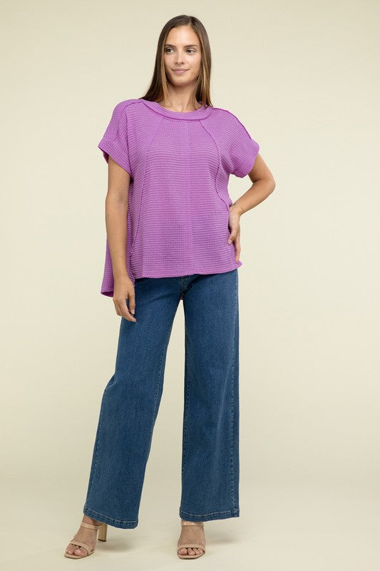 A woman with long brown hair is wearing a purple Brushed Waffle Exposed-Seam Short Sleeve Top and blue jeans, standing against a plain beige background. This casual wardrobe choice, with its short sleeves and side slits, is perfect for everyday wear.