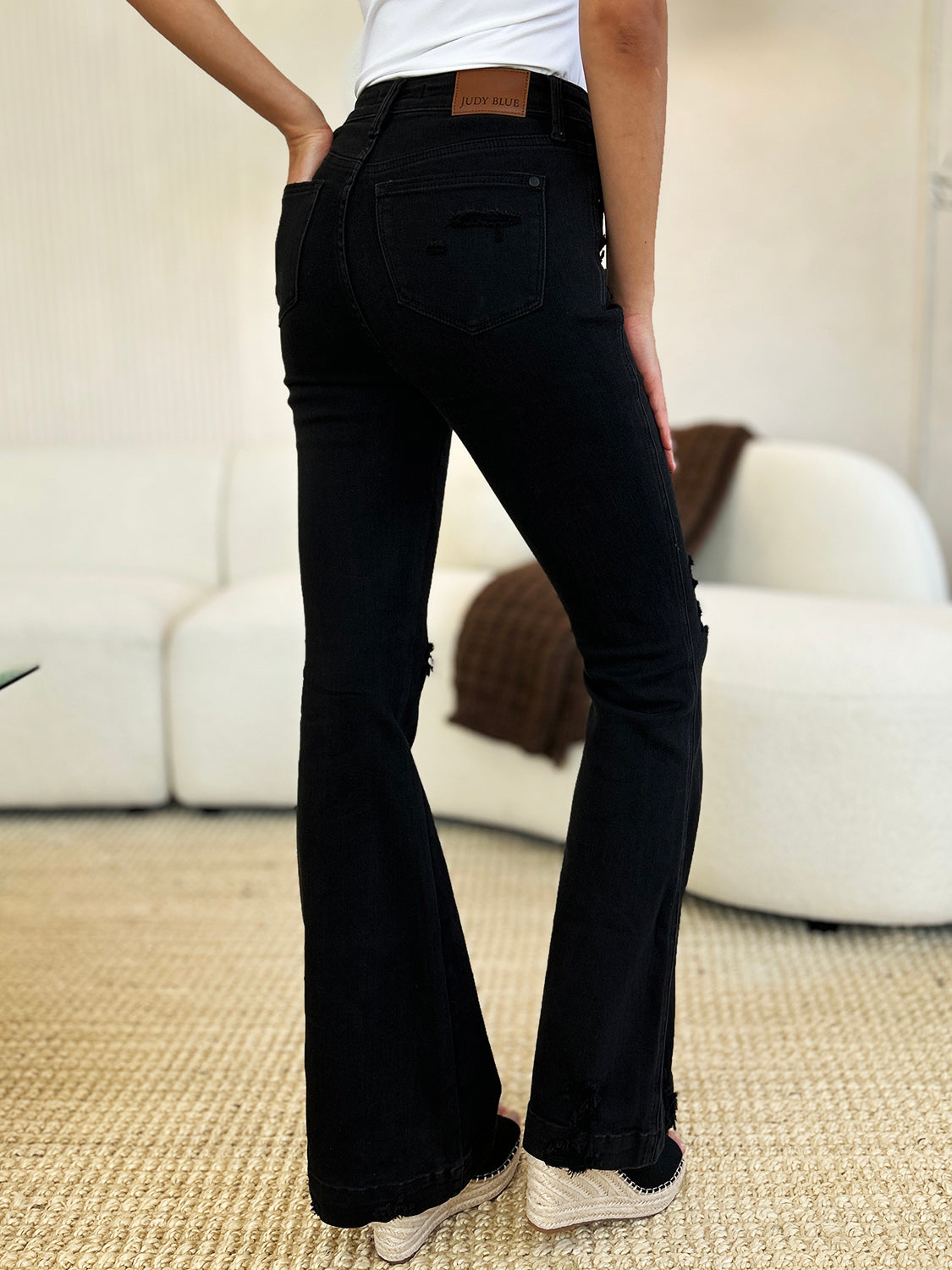 A person stands indoors, hands in pockets, wearing a white top and the Judy Blue Full Size High Waist Distressed Flare Jeans that create a flattering silhouette. In the background, a beige sofa and potted plants add to the retro-inspired look.