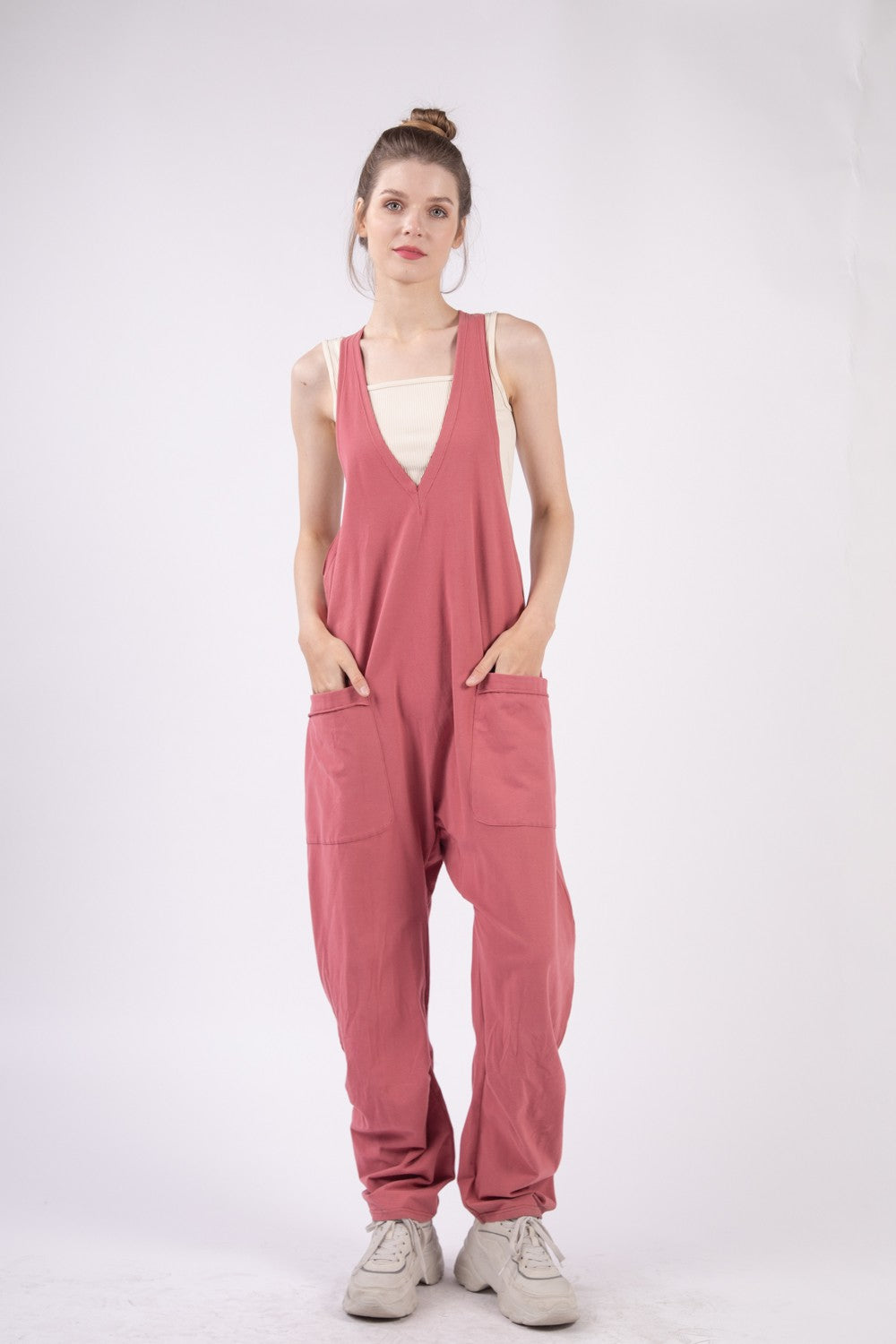 Someone is standing against a plain backdrop, wearing the VERY J Plunge Sleeveless Jumpsuit in pink and white shoes, with their hands casually tucked into the jumpsuit's pockets.