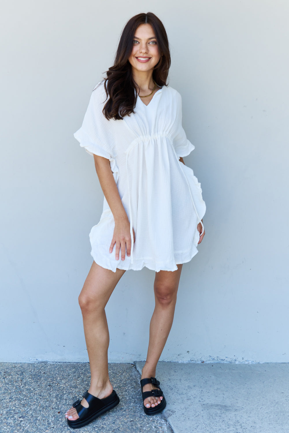 A person with long dark hair smiles while posing in the Ninexis Out Of Time Full Size Ruffle Hem Dress with Drawstring Waistband in White against a plain background.