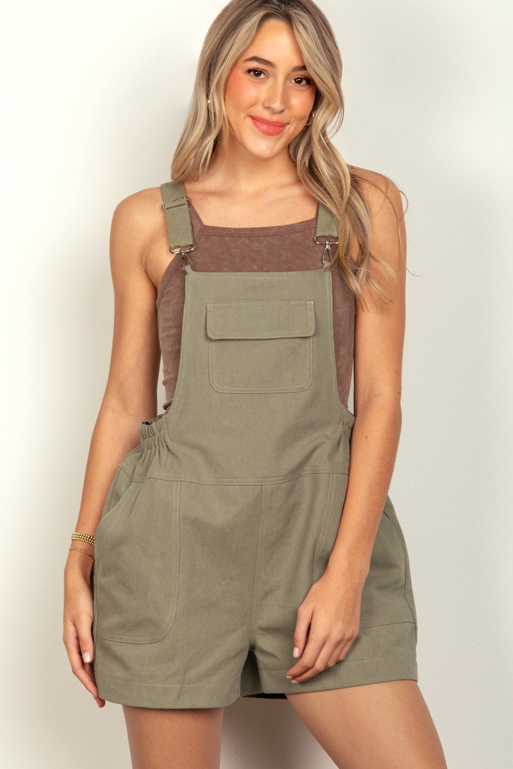 A woman with long blonde hair is wearing a pair of olive green VERY J Adjustable Suspender Overalls with an elastic waist, while her hands rest in the front pockets.