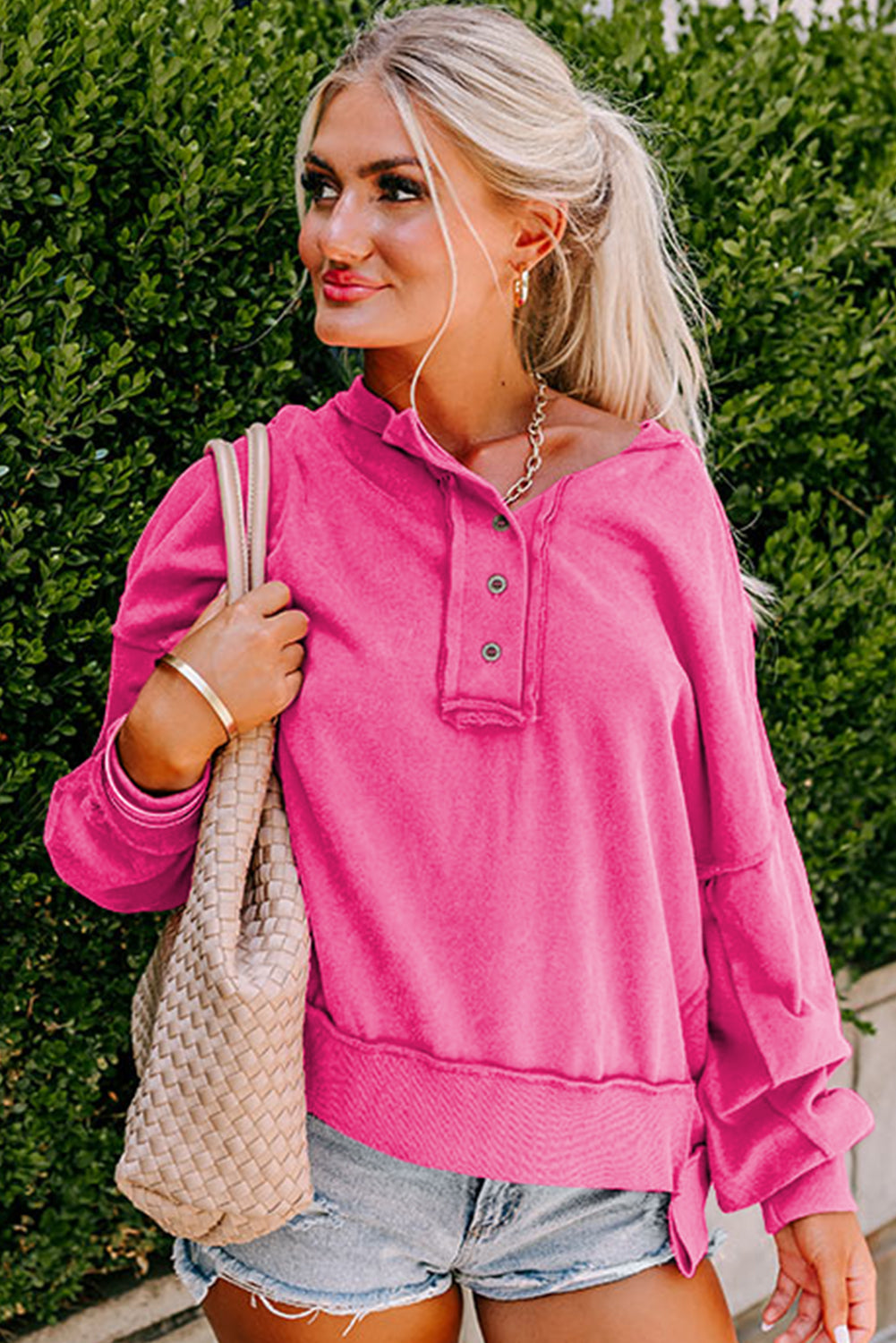 A person with long blonde hair in a ponytail, wearing a Drop Shoulder Henley Buttons Sweatshirt and denim shorts, is standing with their back to the camera in front of a green shrub.