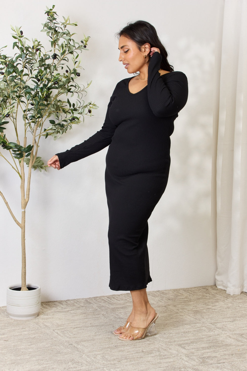 A person stands indoors wearing the chic Culture Code Full Size Ribbed Long Sleeve Midi Slit Dress paired with sandals, next to a potted plant.