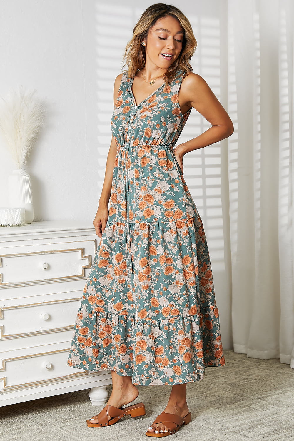 A woman in a Perfee Floral V-Neck Tiered Sleeveless Dress stands indoors near a white dresser. She is wearing brown sandals and smiling.