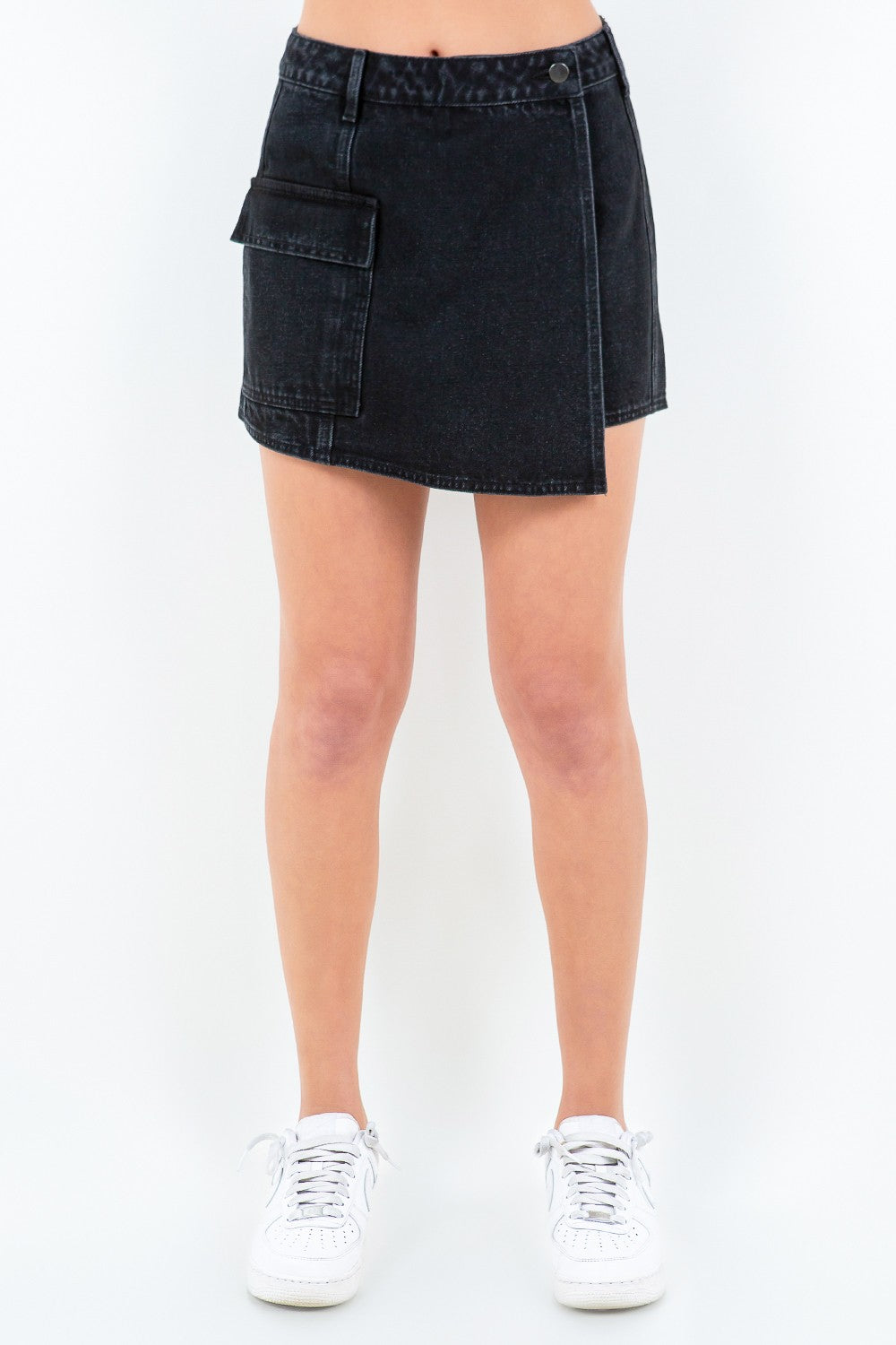 Front view of a person wearing an American Bazi Cargo Pocket Mini Skort in black denim with an asymmetrical front and button waist closure, paired with white sneakers.