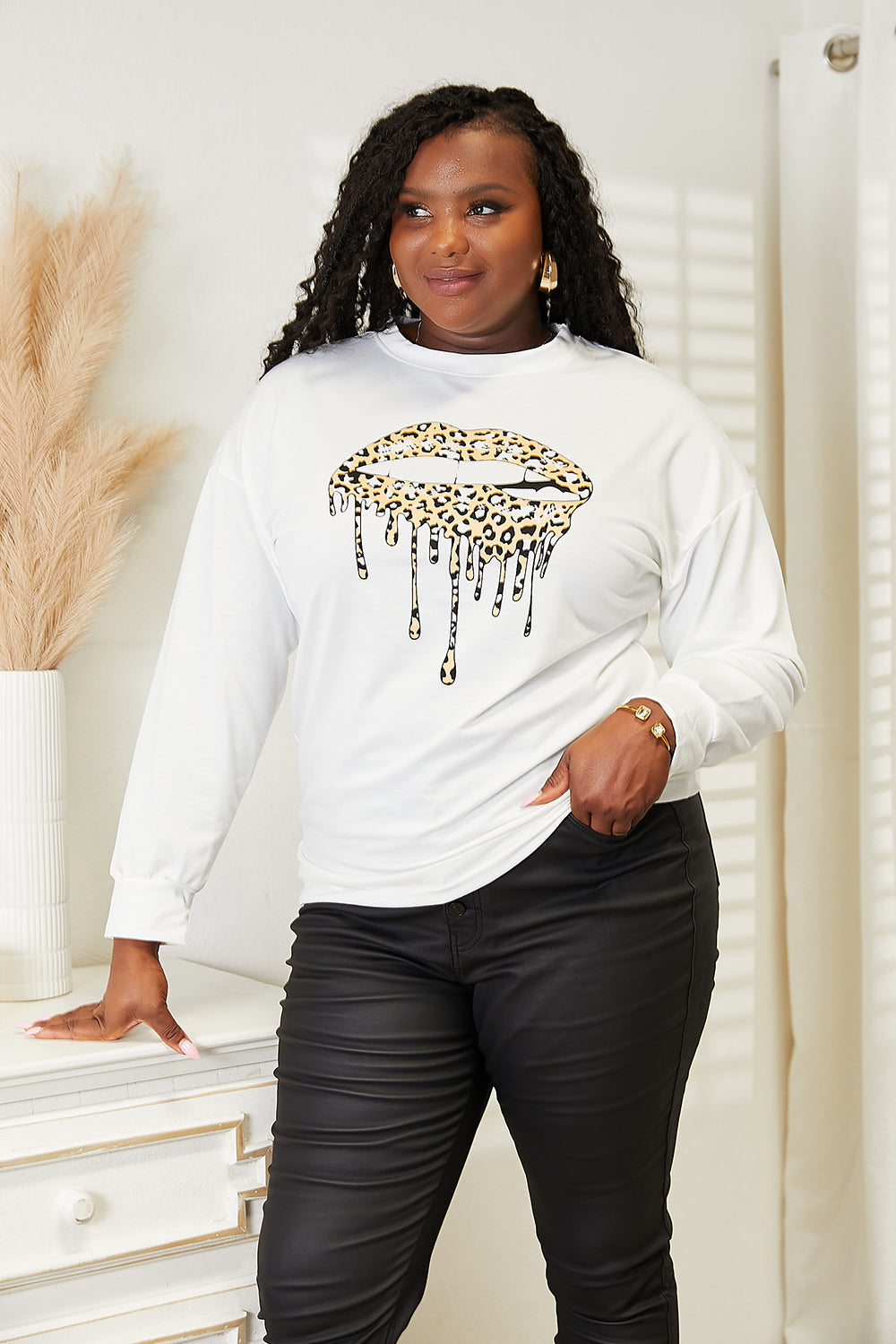 A woman stands indoors wearing a trendy and comfortable outfit—a white Simply Love Graphic Dropped Shoulder Round Neck Sweatshirt, featuring a dripping lip design, paired with black pants. One hand rests on a piece of furniture.