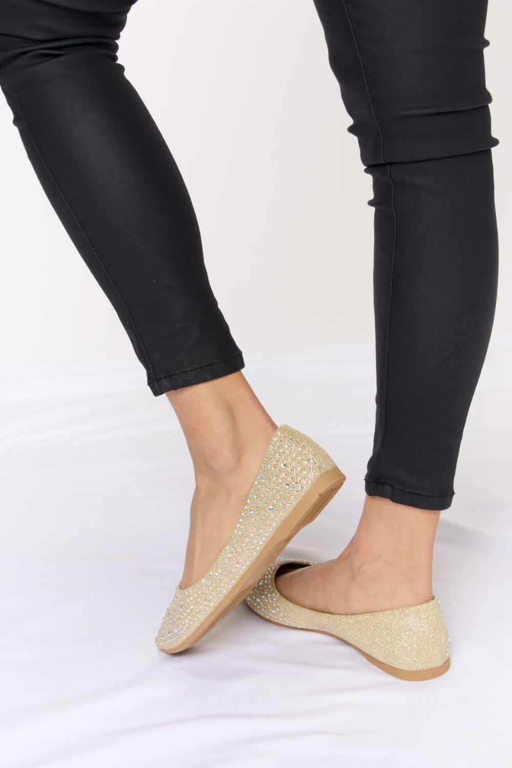 Someone is sitting on a white surface wearing black pants and chic Forever Link Rhinestone Point Toe Flat Slip-Ons in sparkling gold.