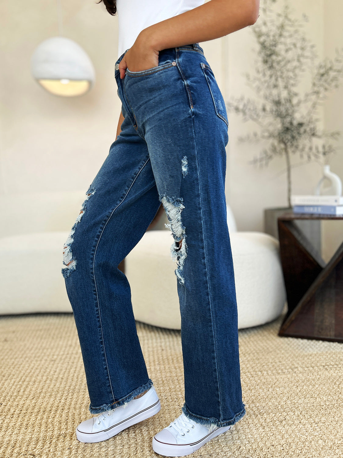 Person standing indoors wearing Judy Blue Full Size Mid Rise Distressed Raw Hem Jeans and black sneakers.