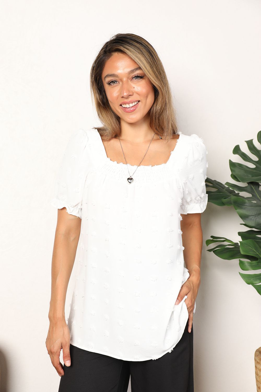 A person wearing the Mandy Swiss Dot Puff Sleeve Square Neck Blouse in white, paired with black pants, stands in front of a light background with green leaves visible on the right side.