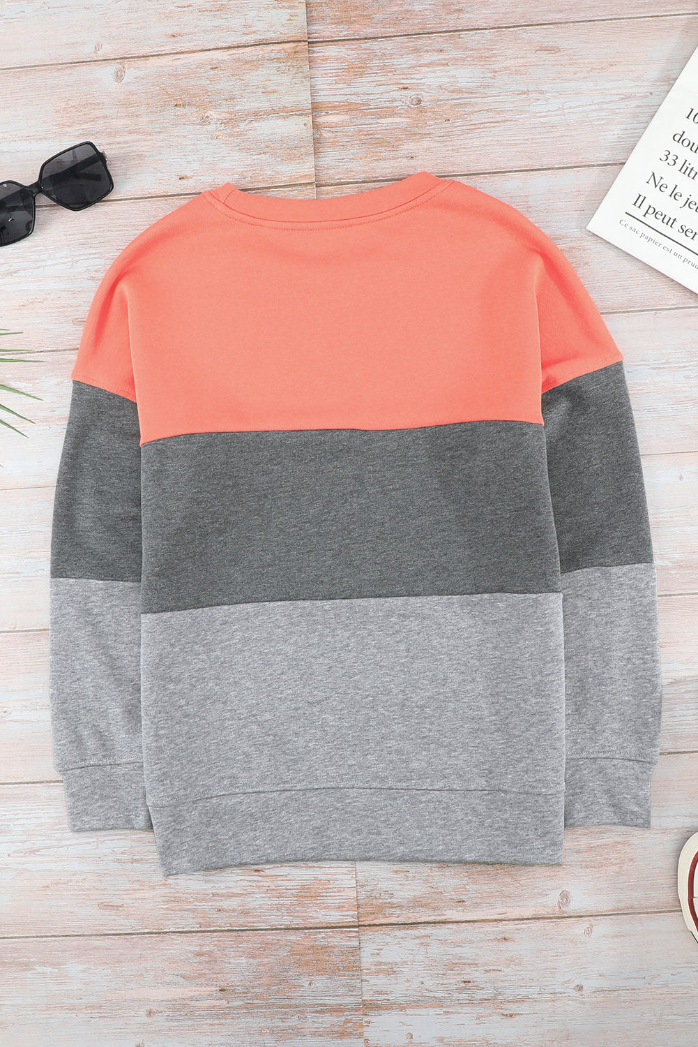 A Colorblock Gray Contrast Stitching Sweatshirt with Slits, made of comfy fabric and featuring coral, gray, and light gray sections, is placed on a wooden surface near sunglasses, a book, and some plant leaves.