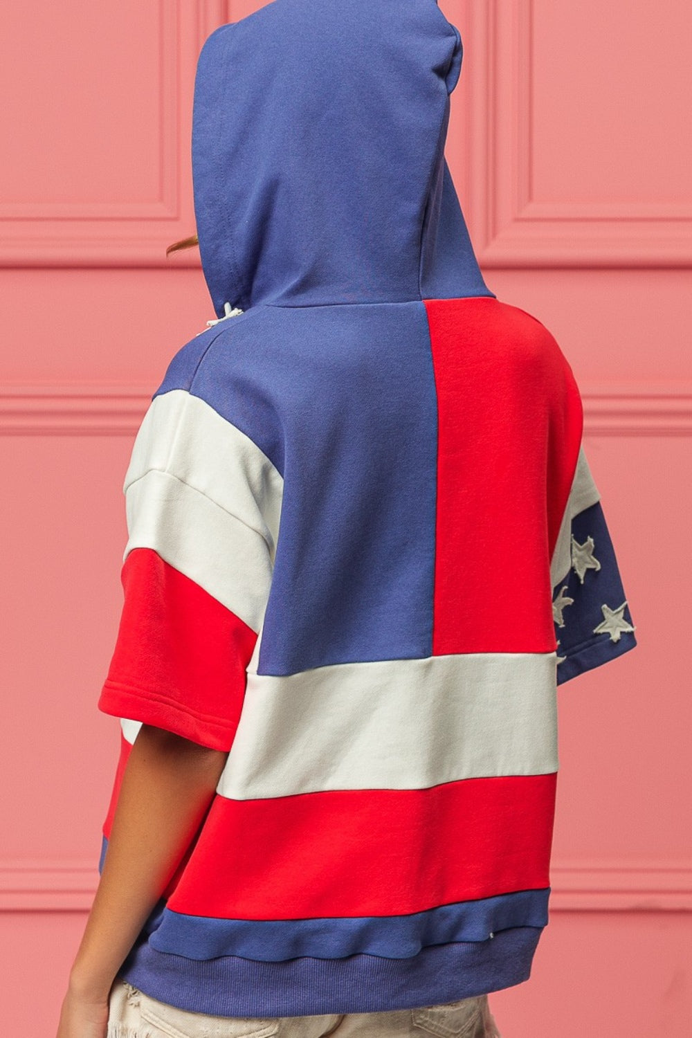 Woman wearing a BiBi American Flag Theme Hoodie, standing against a pink background.