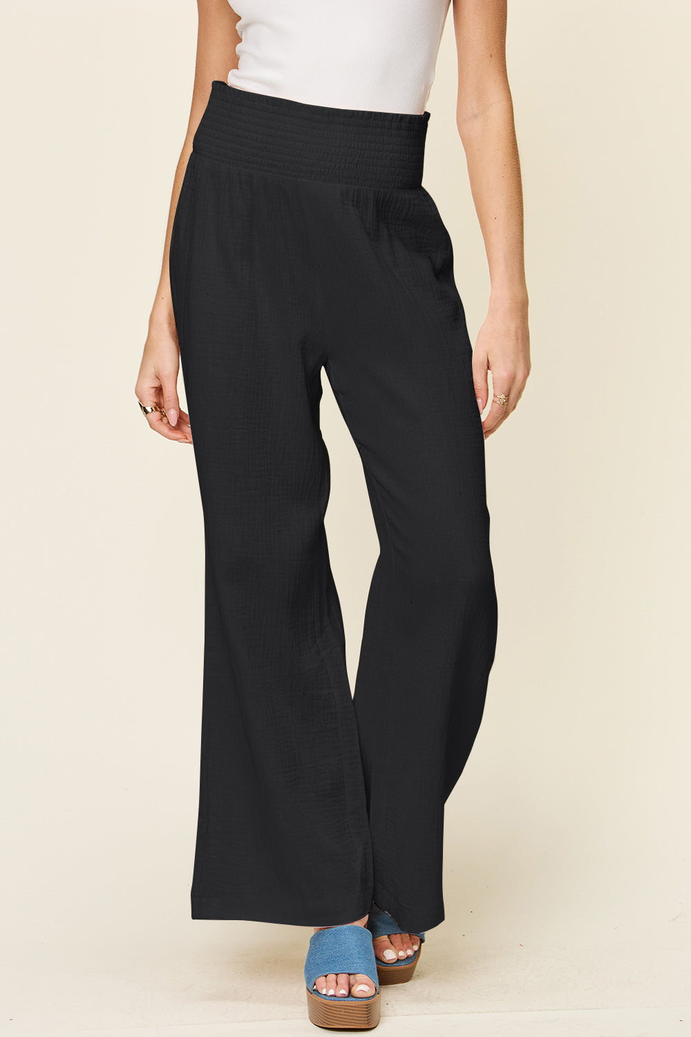 A person wearing Double Take Full Size Texture Smocked Waist Wide Leg Pants in black and a white 100% cotton top is standing with one hand on their hip. They are also wearing blue open-toe sandals.