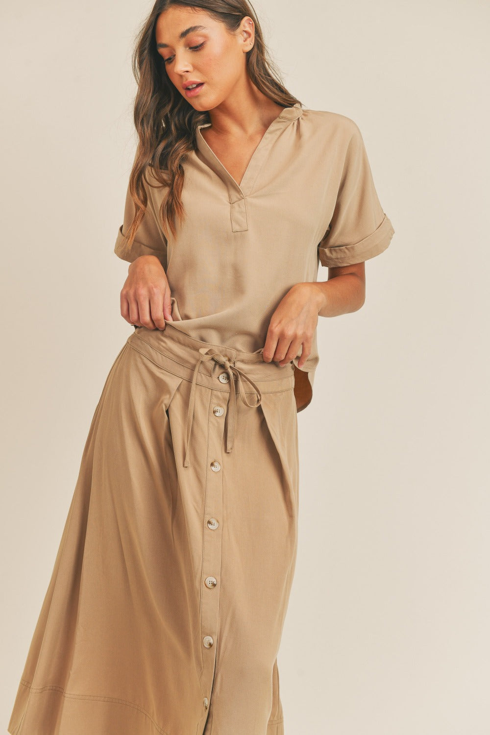 A woman is standing against a plain background wearing the MABLE Short Sleeve Top and Button Down Midi Skirt Set in beige. The elegant two-piece set, which includes a belted midi skirt, is paired perfectly with beige heels as she confidently poses with one hand on her hip.