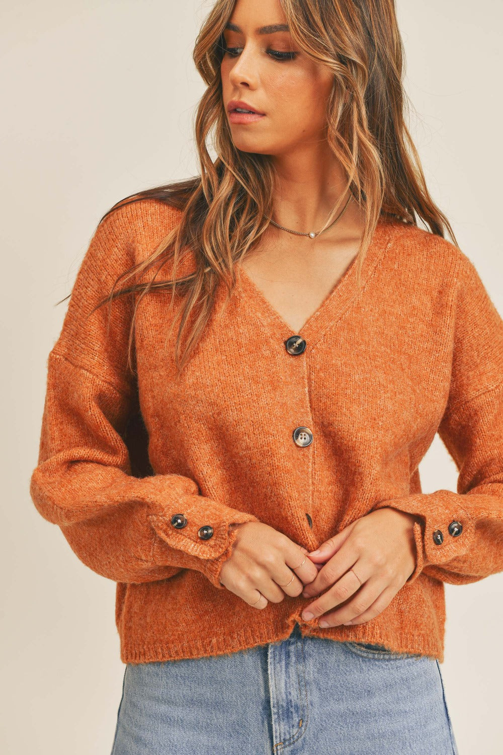 A woman wearing a MABLE Long Sleeve Button Down Sweater Cardigan in burnt orange and light blue jeans looks to her left against a plain background, exuding classic elegance.