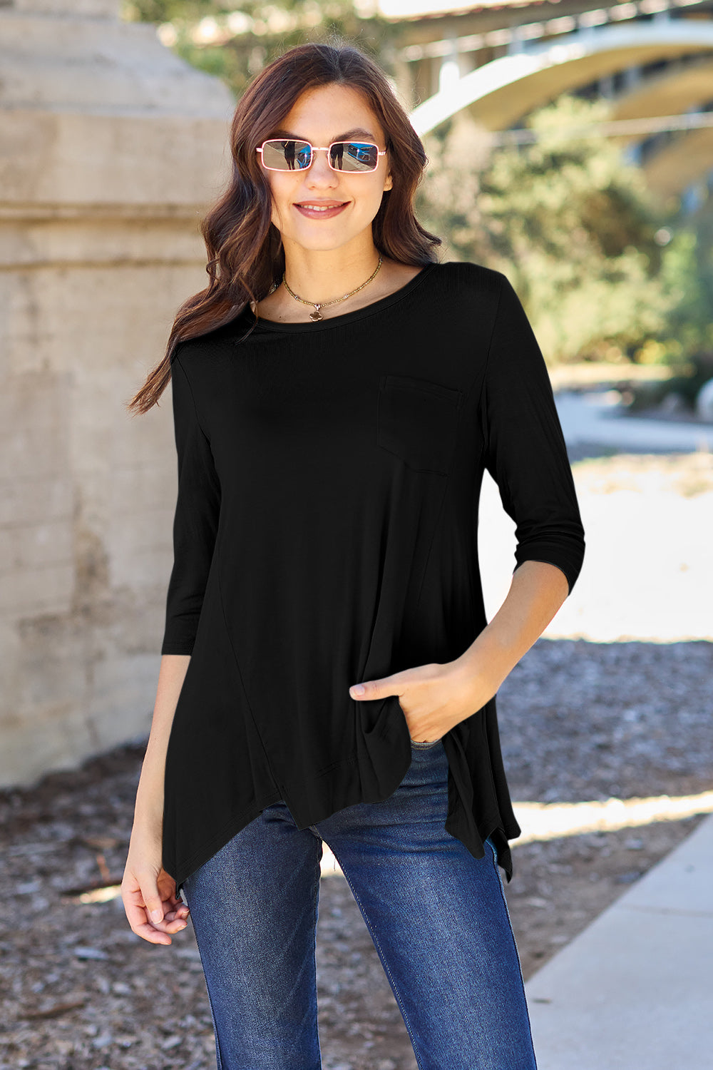 A woman stands outdoors wearing sunglasses, a Basic Bae Full Size Round Neck Pocketed T-Shirt with an asymmetrical hem and chest pocket made of rayon spandex, and blue jeans. She is smiling and has one hand in her pocket. Her basic style radiates effortless charm.