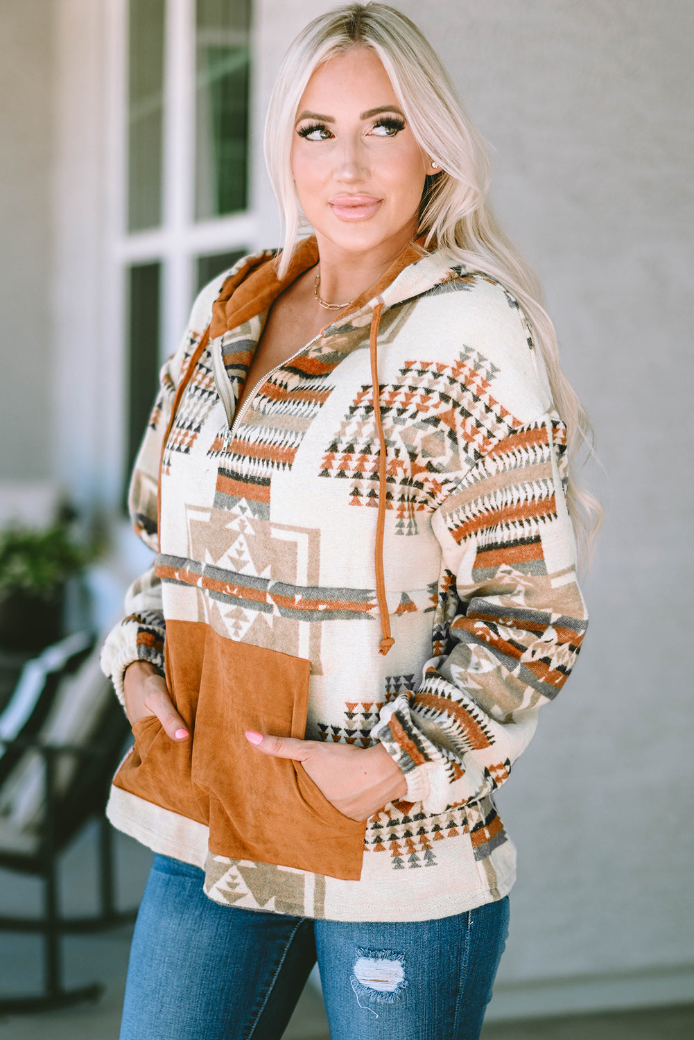 A person with long blonde hair, seen from behind, is wearing the Beige Aztec Print Kangaroo Pocket Half-Zip Hoodie. The hoodie showcases an Aztec pattern in shades of brown, beige, and orange and features a drop shoulder design with a cozy kangaroo pocket, all while standing outdoors.