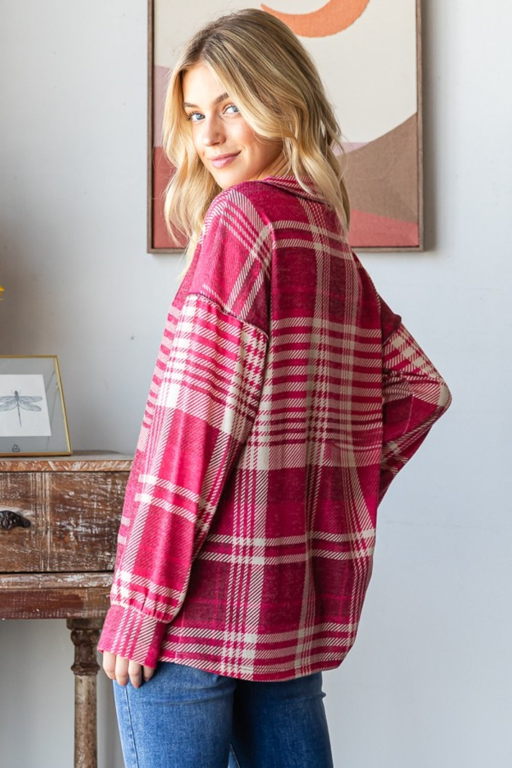 A person with long blond hair smiles while wearing the Heimish Plaid Drop Shoulder V-Neck T-Shirt in red and white and a pair of blue jeans, leaning against a wooden table with framed pictures in the background.