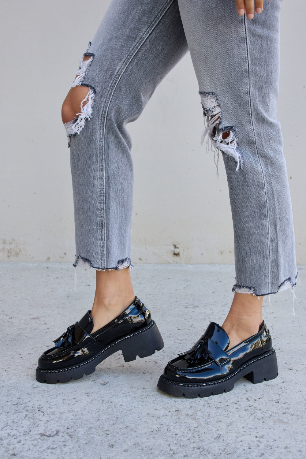 A person wearing ripped gray jeans and the Forever Link Tassel Detail Mid Heel Chunky Loafers, which feature shiny black tones, chunky soles, and a subtle tassel detail.