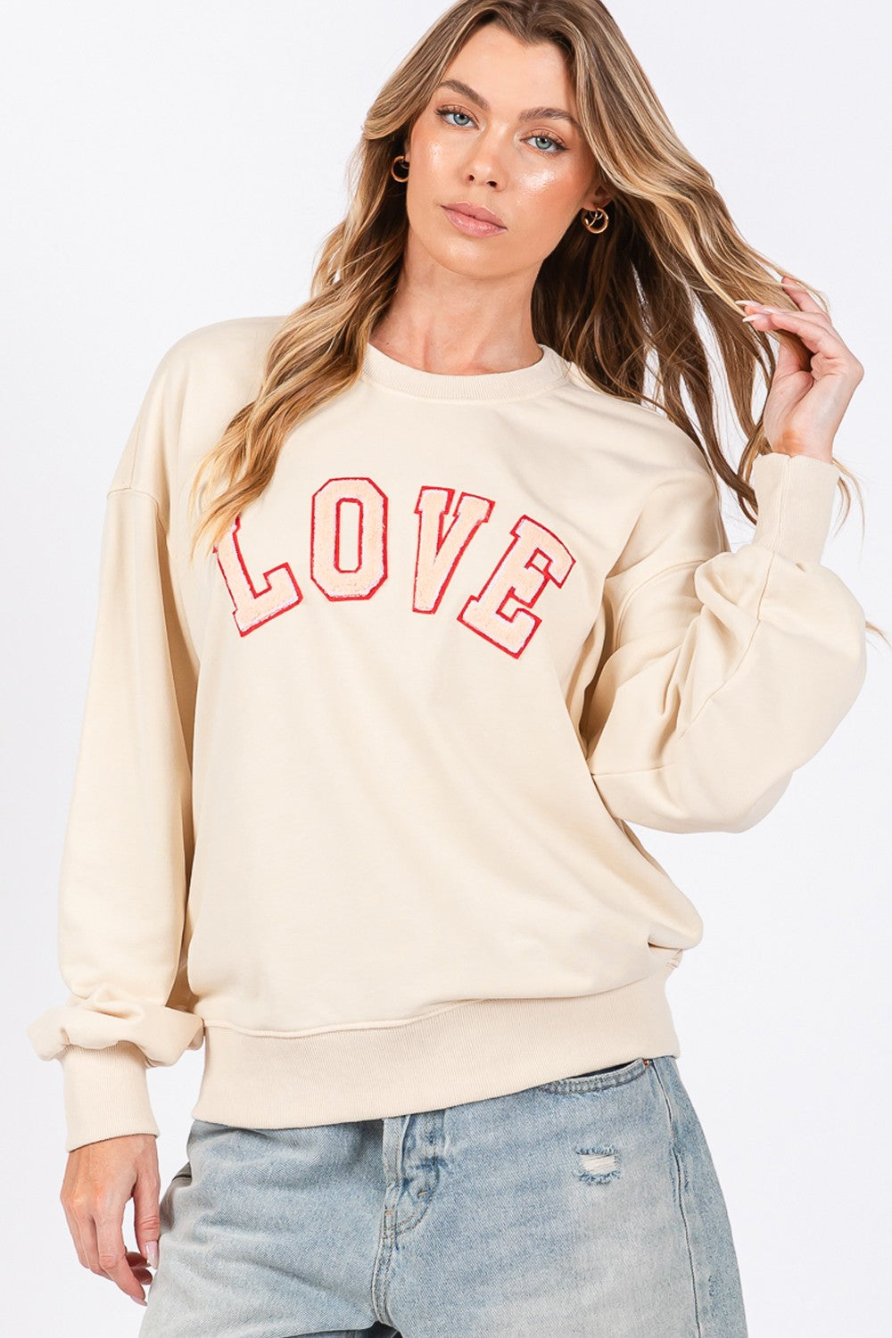 A woman dressed in a SAGE + FIG LOVE Path Applique Drop Shoulder Sweatshirt and a wide-brimmed hat is touching the brim of her hat with both hands, looking to the side and flaunting her cozy and stylish sweatshirt.