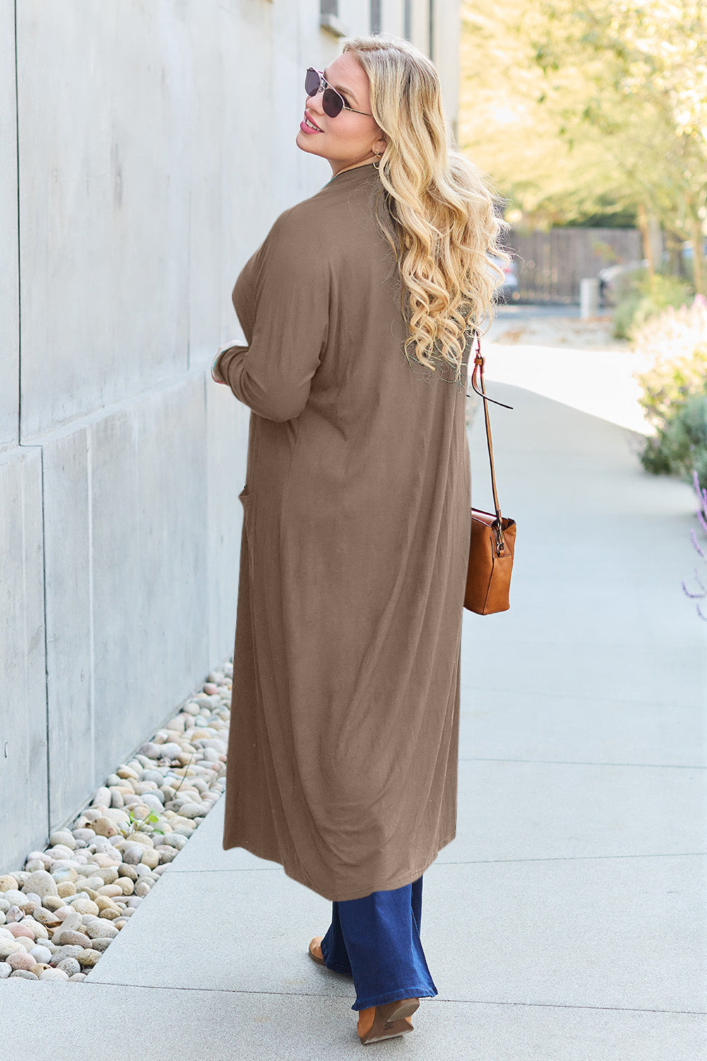 A woman stands against a concrete wall, wearing the Basic Bae Full Size Open Front Long Sleeve Cover Up made from rayon spandex, a white top, blue jeans in her perfect size, and brown shoes with her hands in her pockets. Machine wash cold for best results.