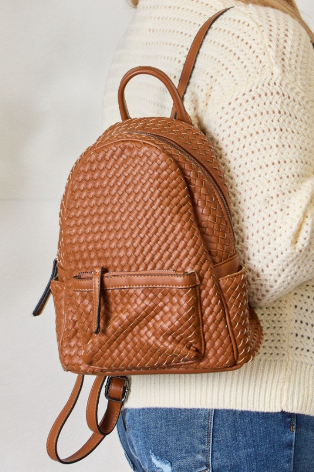 A person wearing a cream sweater and jeans is carrying the SHOMICO PU Leather Woven Backpack in brown.