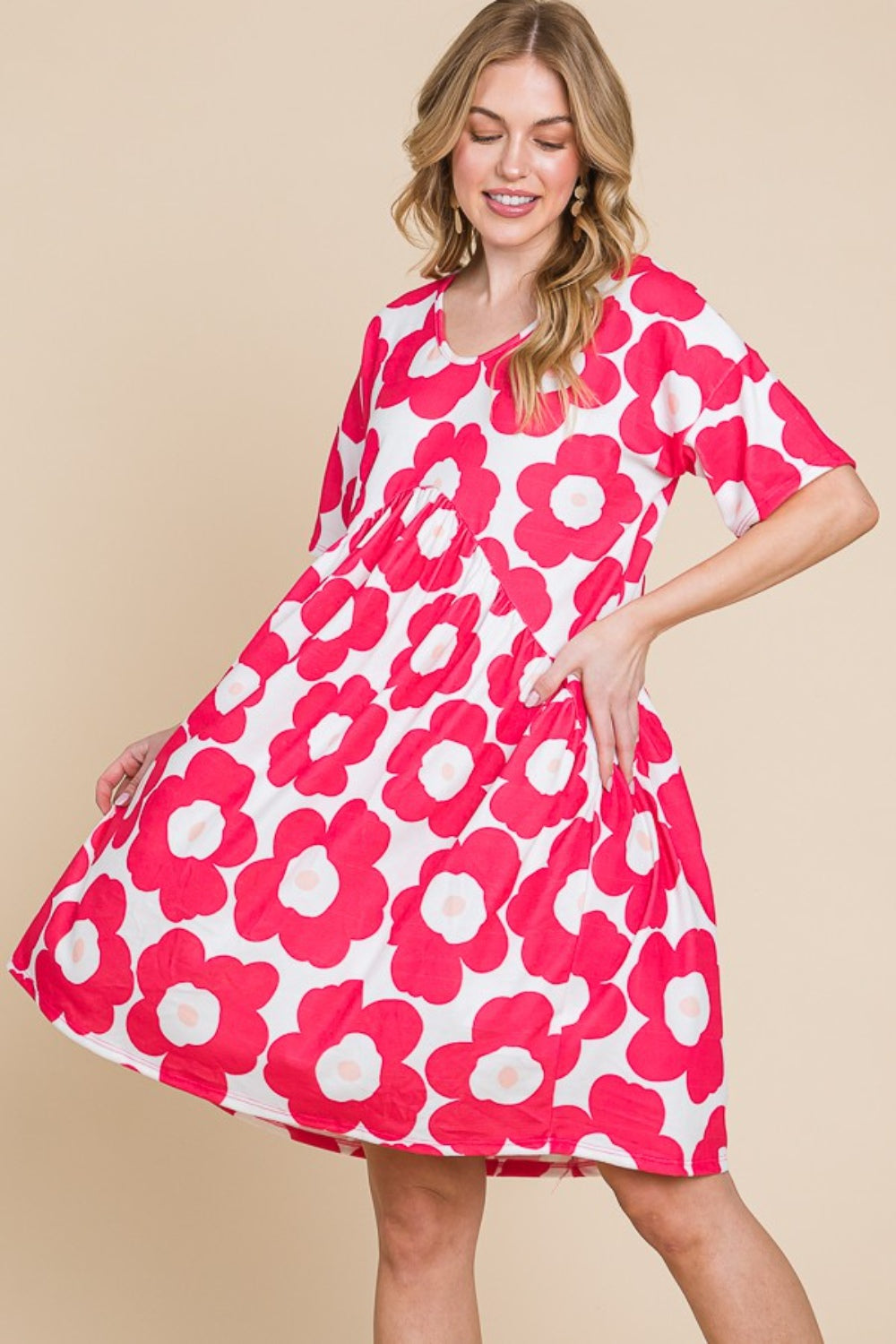 A woman gracefully stands against a beige background, wearing the BOMBOM Flower Print Ruched Dress adorned with large pink blooms. The stretchy fabric of the dress complements her form, effortlessly enhancing its elegant silhouette.