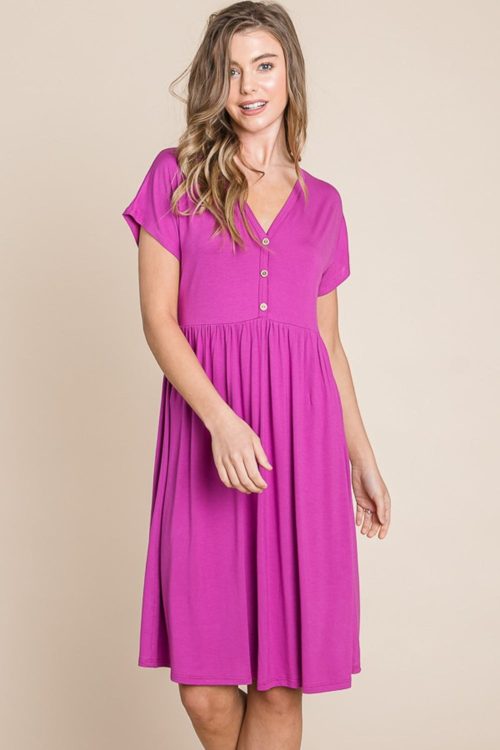 A person stands against a neutral background wearing the BOMBOM V-Neck Short Sleeve Dress, featuring a vibrant pink color and button details, crafted from soft rayon spandex fabric.