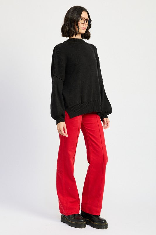 A woman wearing an OVER FITTED LONG SLEEVE SWEATER TOP made of 60% cotton in brown and khaki shorts holds a denim handbag with a brown strap against a neutral background.