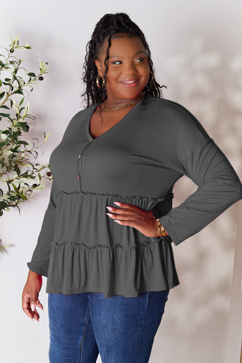 Person stands indoors near a plant, wearing the Double Take Half Button Long Sleeve Ruffle Hem Blouse in gray and blue jeans, smiling while looking to the side. The slightly stretchy fabric of the blouse adds comfort while its buttoned design offers a touch of elegance.