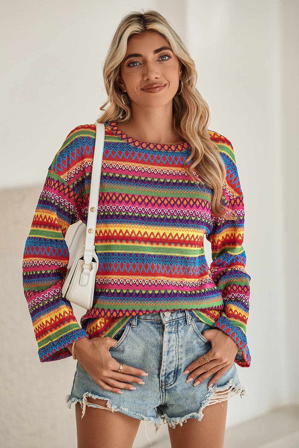 A person stands with hands in the pockets of their denim shorts, wearing a colorful Rose Red Ethnic Wavy Pattern Drop Shoulder Knit Top and a white shoulder bag.