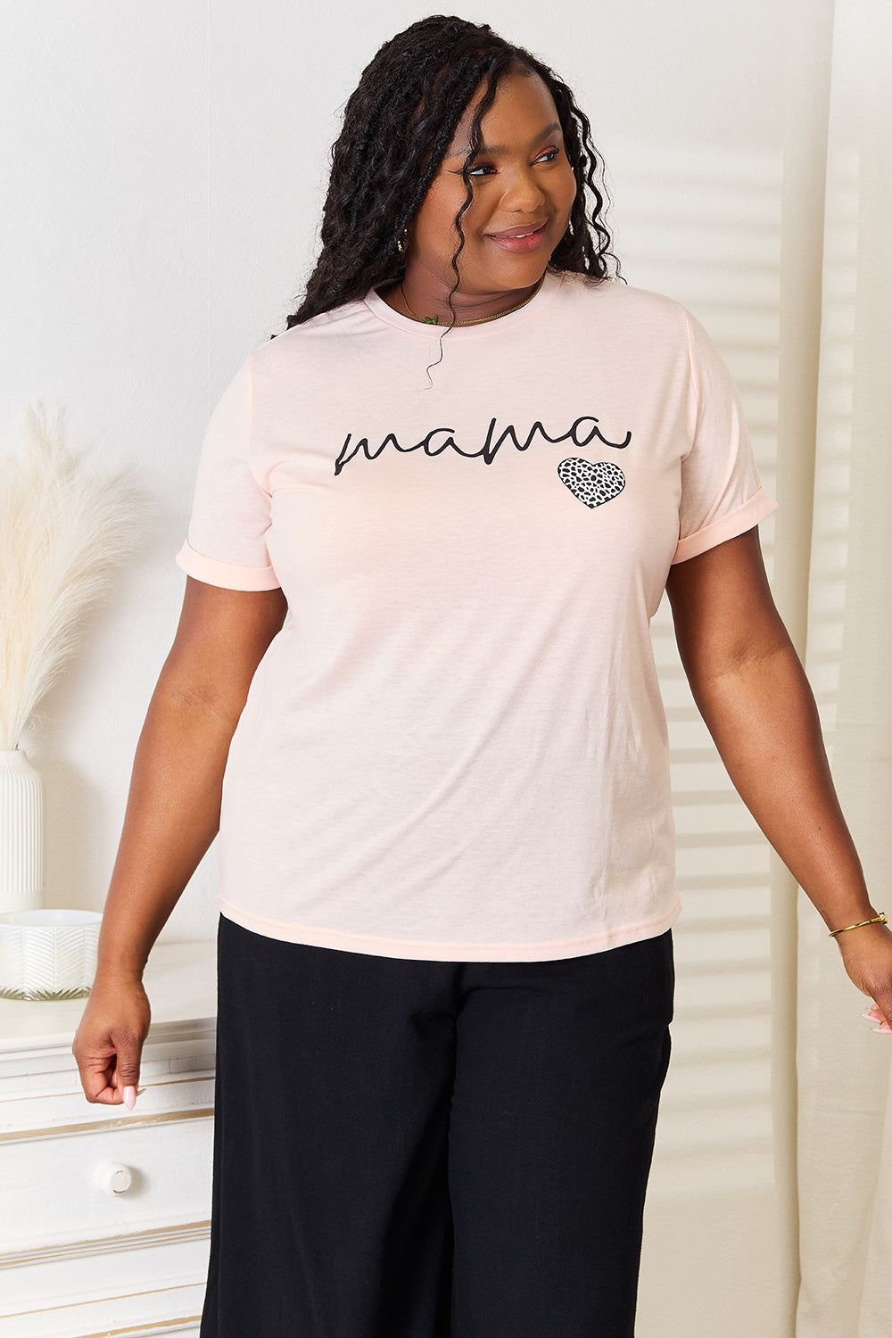 A woman with long dark hair, wearing a light pink Simply Love MAMA Heart Graphic T-Shirt and high-waisted black leggings, stands in front of a white dresser. The t-shirt, featuring the word "mama" and a small heart design, makes for a perfect motherhood gift.