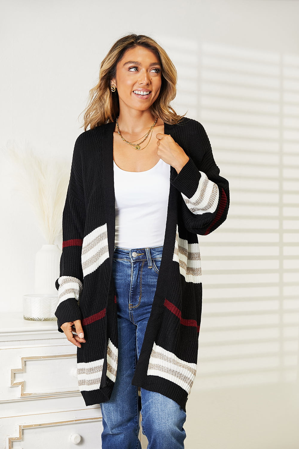 A person wearing a Perfee Striped Rib-Knit Drop Shoulder Open Front Cardigan with white and red accents, a white top, blue jeans, and layered necklaces stands next to a decorative table.