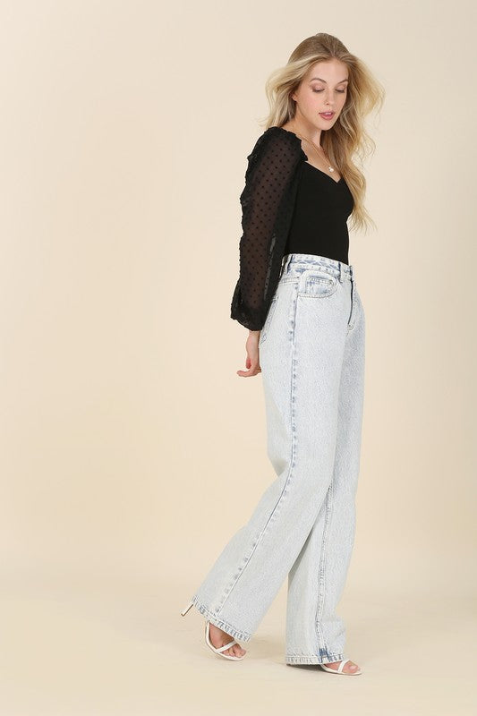 A person wearing the LS shirring sleeve bodysuit with Swiss dot mesh, featuring sheer, long puffed sleeves and a sweetheart neck, paired with light-colored jeans, stands against a plain background.