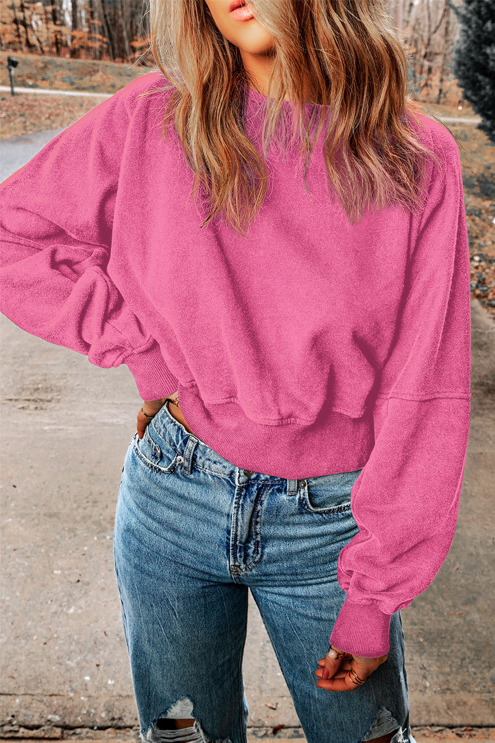 A person with wavy blonde hair is wearing a Rose Acid Wash V-shape Open Back Sweatshirt and blue jeans, walking on a paved path outdoors.