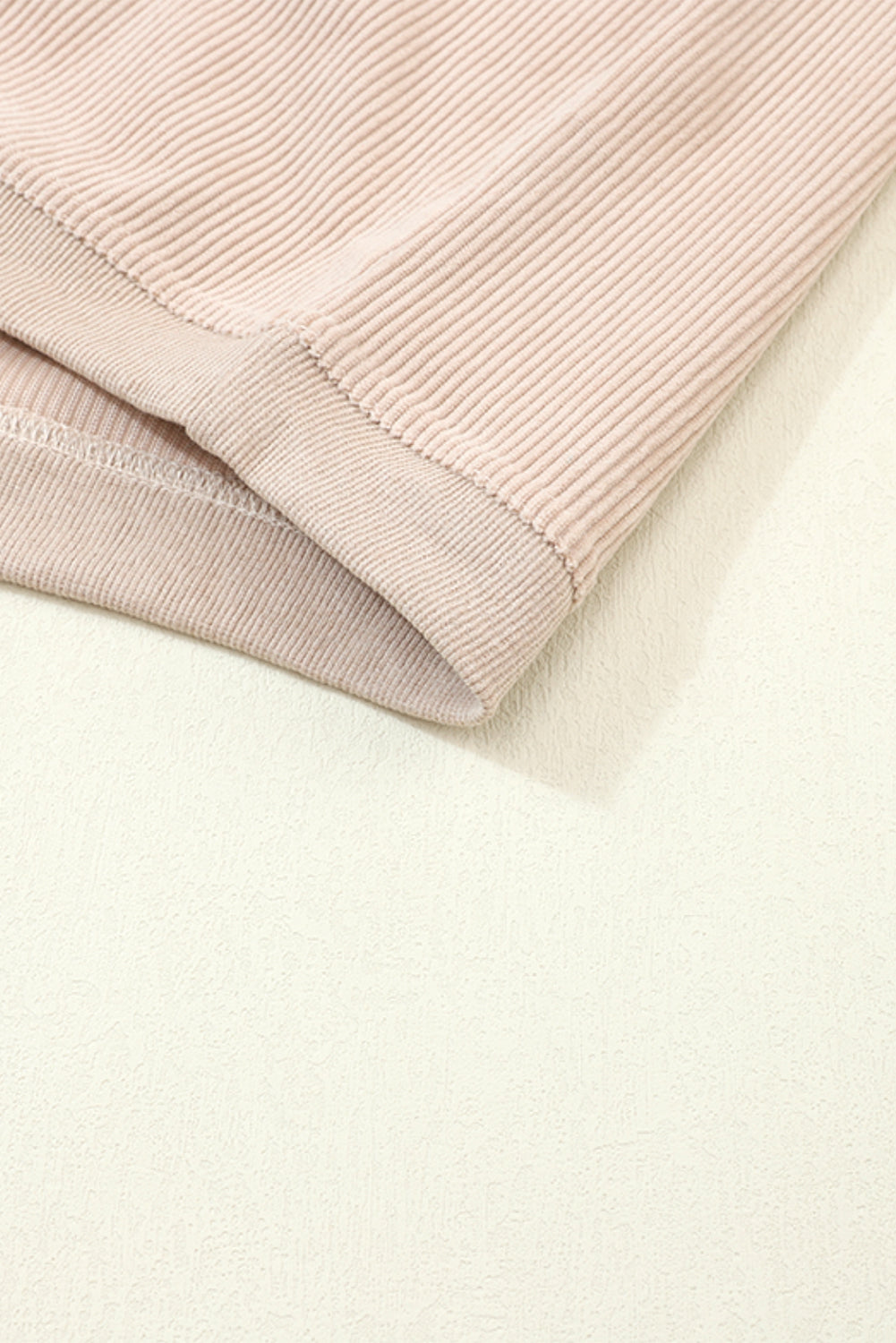 A woman stands outdoors, looking down while touching her hair with one hand. She is wearing an Apricot Ribbed Corduroy Oversized Sweatshirt and white pants, exuding an effortlessly chic and fashionable look.