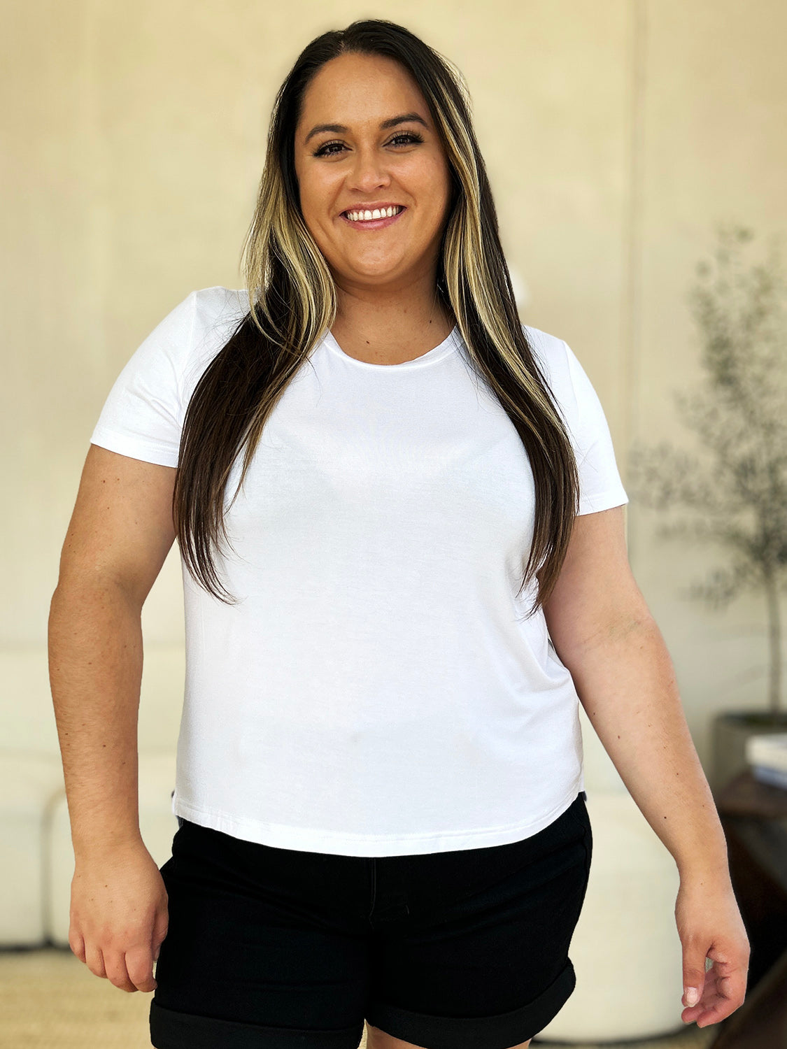 A woman stands next to an indoor plant, smiling, with one hand in her khaki pants pocket. In her pink Basic Bae Full Size Round Neck Short Sleeve T-Shirt and gold jewelry, she radiates casual elegance.