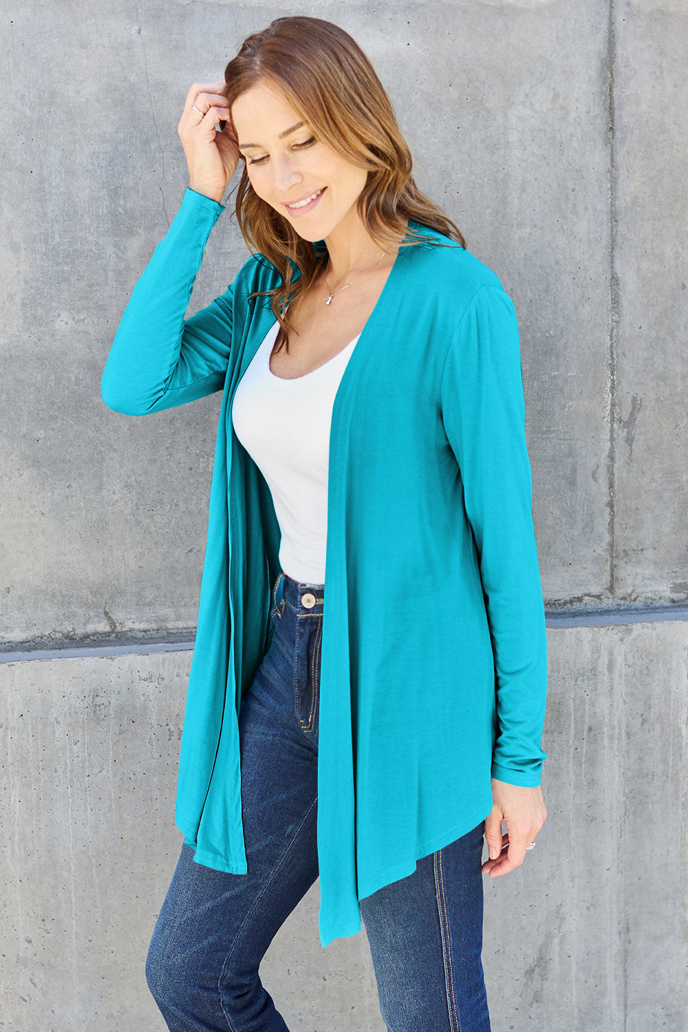 A woman wearing the Basic Bae Full Size Open Front Long Sleeve Cardigan in navy blue, paired with a white top and blue jeans, stands against a concrete wall. She touches her hair and gazes to the side, embodying a chic yet simple style. The slightly stretchy materials of the cardigan ensure comfort while maintaining its stylish simplicity. For easy care, machine wash it cold.