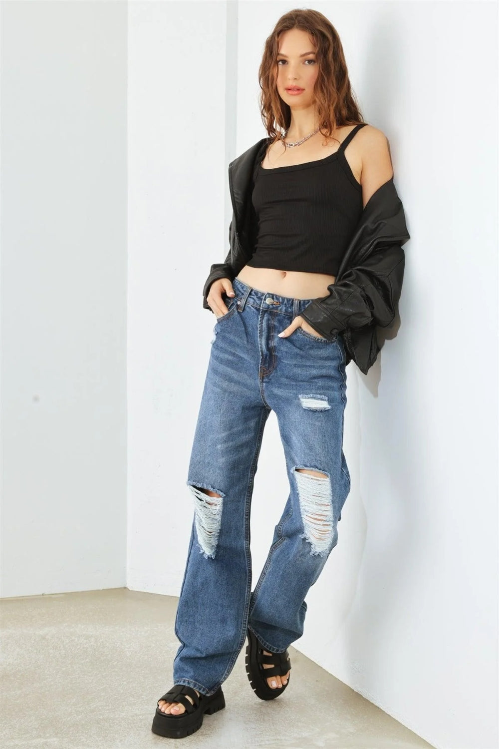 Person wearing HAMMER COLLECTION Distressed High Waist Jeans with ripped knees and black platform sandals, standing in a minimalist setting. The flattering fit and edgy distressed details add to the overall chic look.