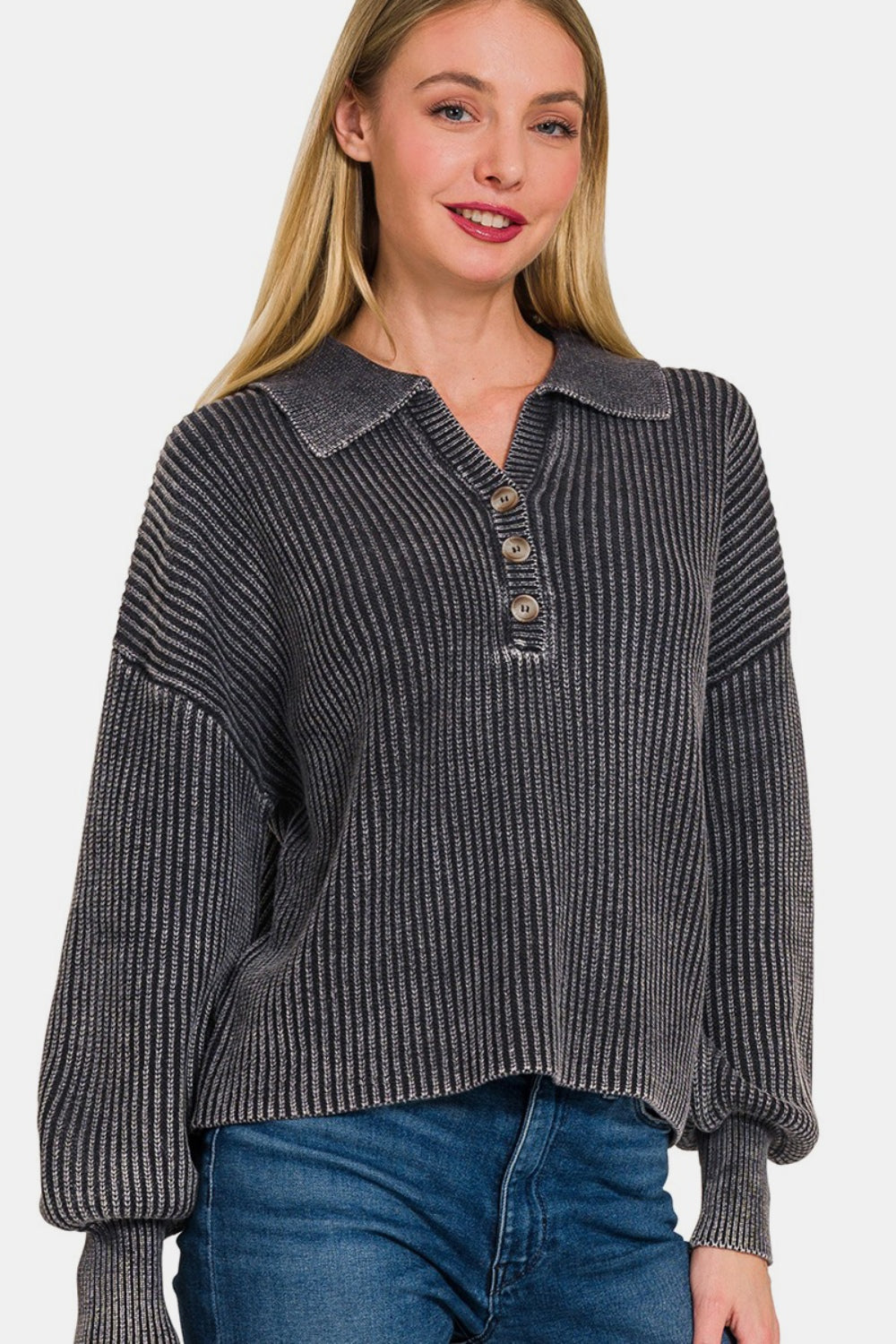 A woman with long blonde hair modeling the Zenana Washed Half Button Long Sleeve Sweater, made of soft washed fabric in grey, paired with blue jeans against a plain backdrop.