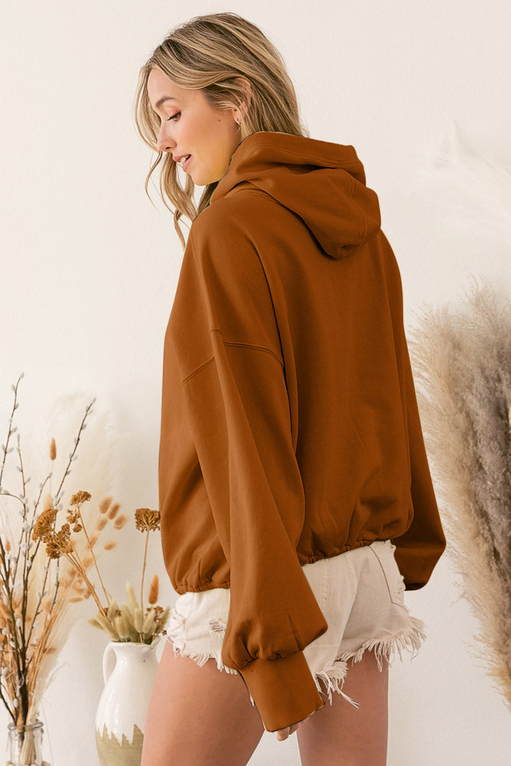 A person wearing a Brown Ribbed Trim Kangaroo Pocket Zipped Hoodie and frayed shorts stands sideways near decorative dried plants in vases.