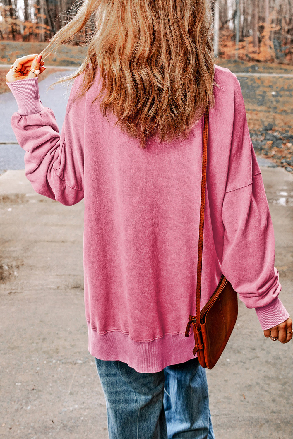 A person with wavy blonde hair, wearing a Pink Drop Shoulder Ribbed Trim Oversized Sweatshirt, blue jeans, and holding a brown bag stands outdoors with their back to the camera.