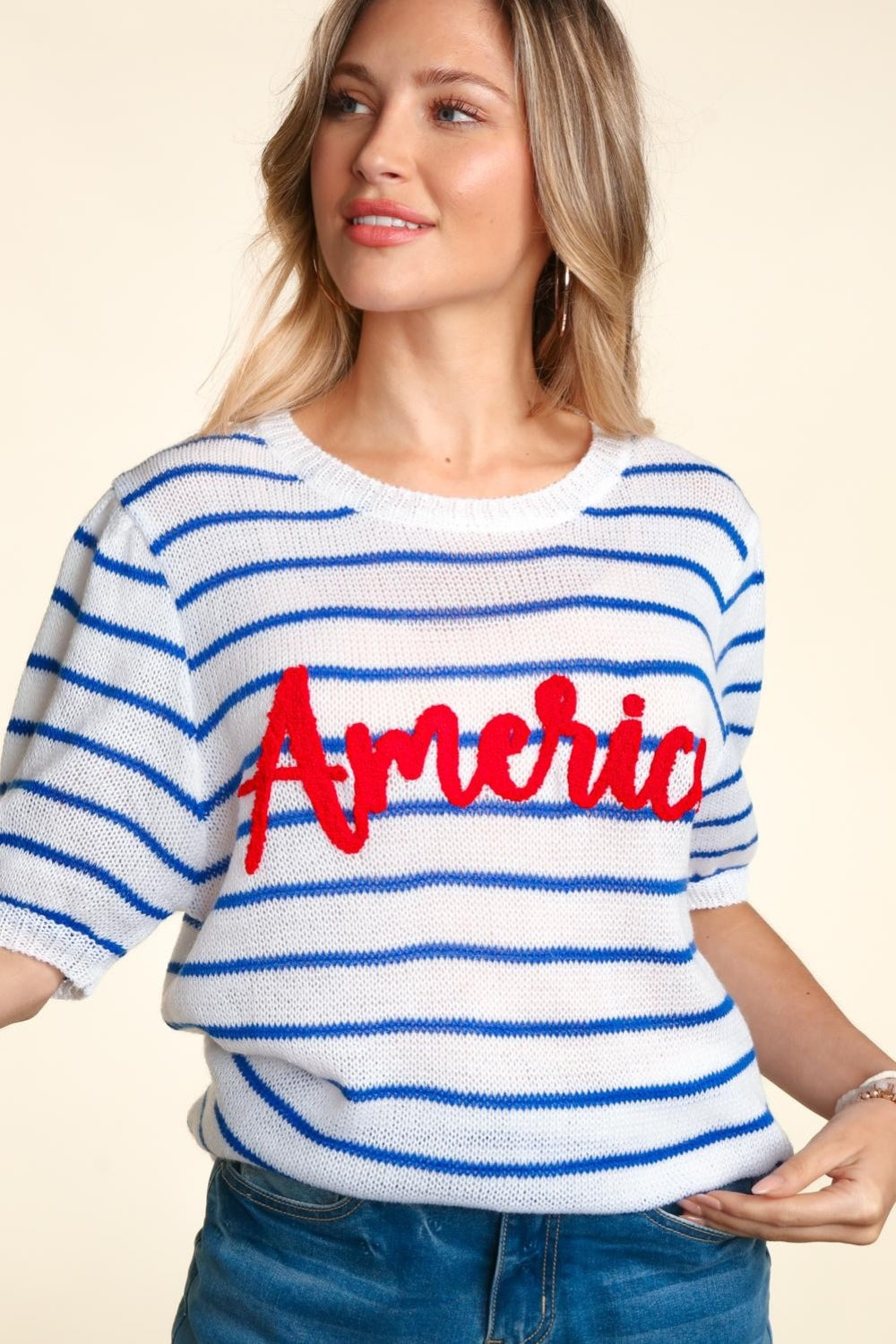 A woman wearing a chic Haptics Letter Embroidery Striped Knit Top featuring "America" in red stands against a neutral background. The white and blue striped knit design pairs perfectly with her denim shorts.