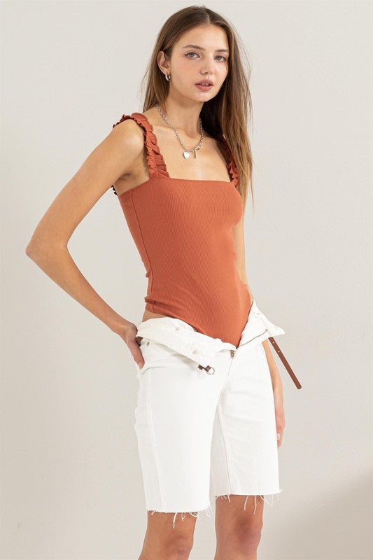 A young woman in a chic Ribbed Ruffle Strap Bodysuit and ripped denim shorts poses against a plain background.