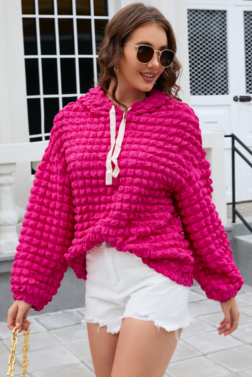 A person stands with their back to the camera, donning the Rose Bubble Textured Waffle Hoodie in a vibrant pink hue. They are in a room with yellow floral wallpaper, and a corner of a wooden chair is visible.