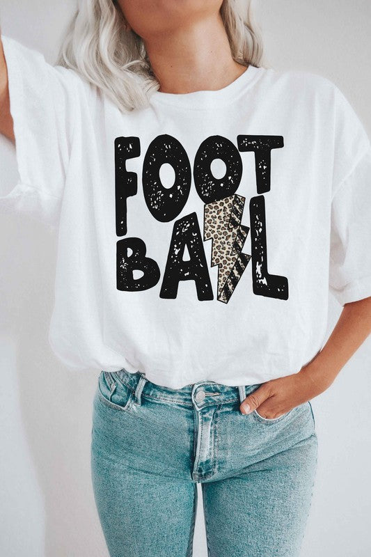 A person wearing a LEOPARD LIGHTNING FOOTBALL Graphic Tee, which features the text "FOOT BALL" and a leopard lightning football design in the center, is standing in a room with a guitar in the background.