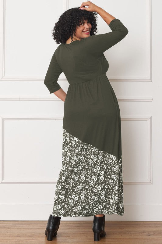 A woman with curly hair is wearing the Asymmetrical Leopard Accent Maxi Dress, featuring a long black design with a floral patterned section at the bottom. She stands against a white paneled wall, her hands tucked in her dress pockets, showcasing its elegant design.