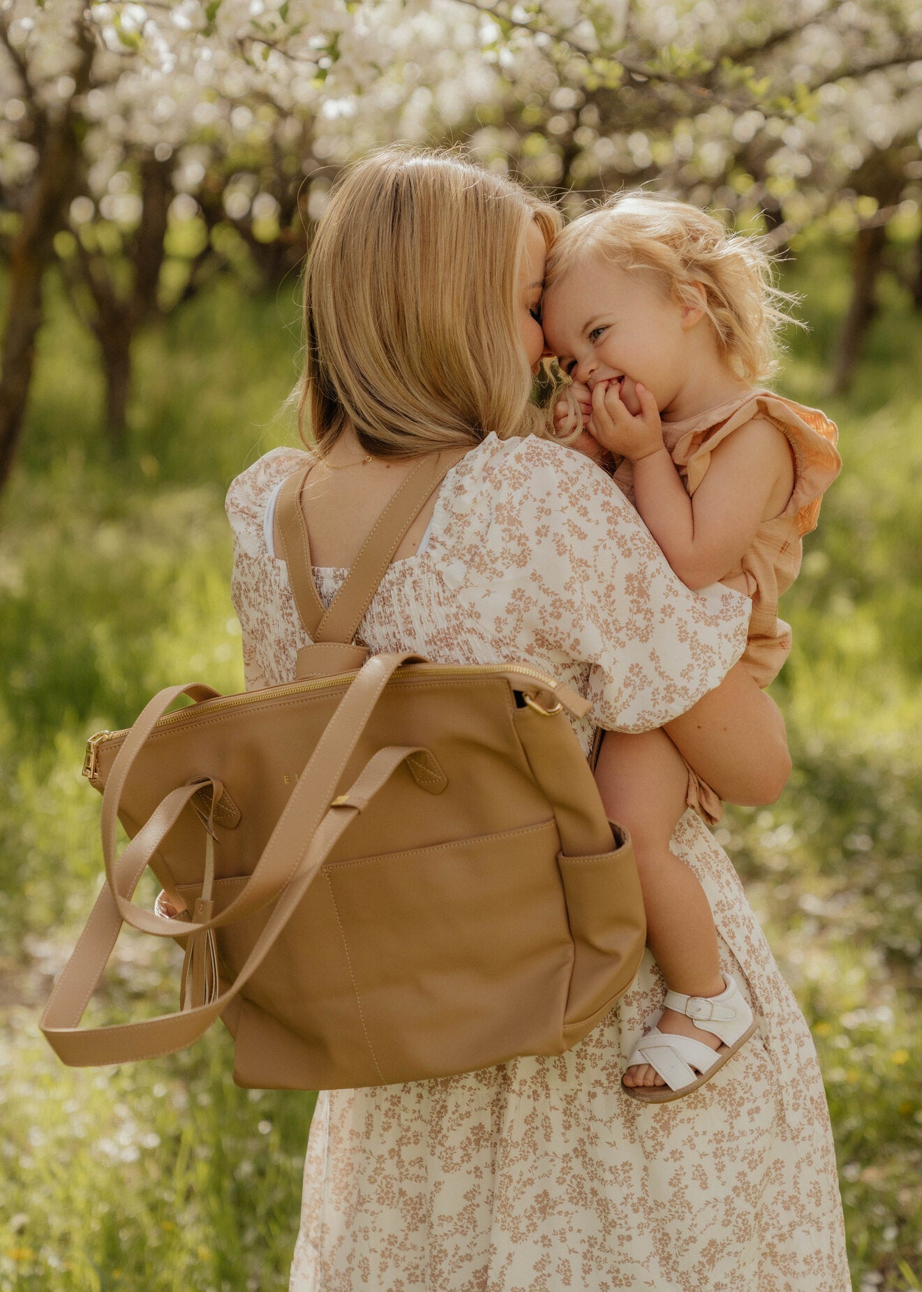 A person stands against a white brick wall, showcasing *The Aberdeen*, a stylish brown vegan leather tote with two front pockets and tassels.